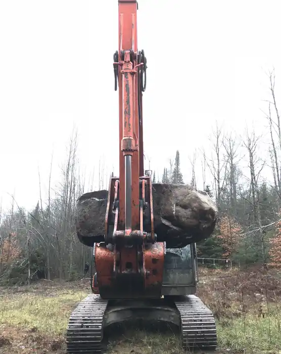 Excavator bucket and thumb gripping a large fieldstone boulder