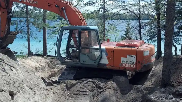 Orange Hitachi EX200 with an Oesch Excavating sticker, digging beside a blue lake
