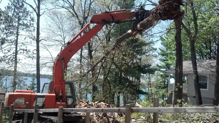 Excavator grapple lifting a felled tree beside a lake cottage